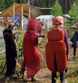 Viisi 12–13-vuotiasta tyttöä metsämaastossa telttojen keskellä. Kolme tyttöä seisoo selkä katsojaan päin muhkeissa, selkäpuolen peittävissä, värikkäissä koppakuoriaisasusteissa. Kaksi tyttöä toimii videokuvaajina tabletin kanssa.